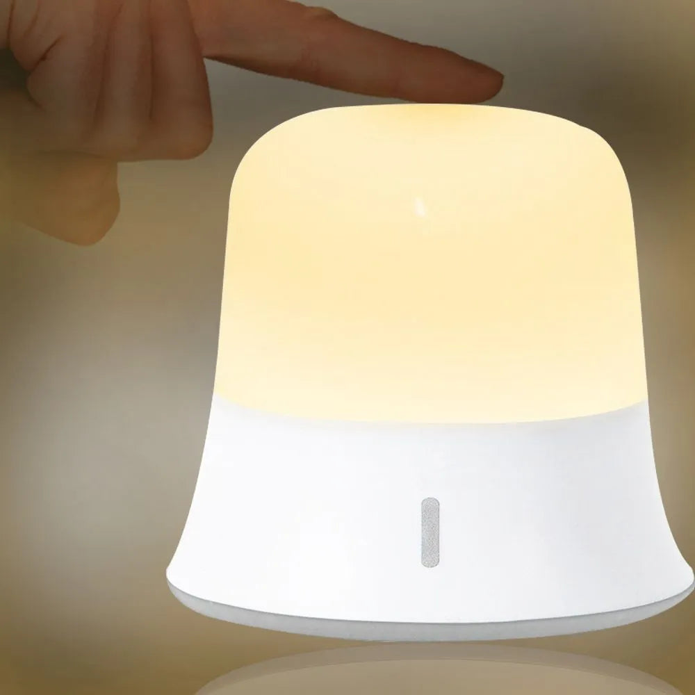 White cylindrical touch-control bedside night light with rechargeable battery on a table.