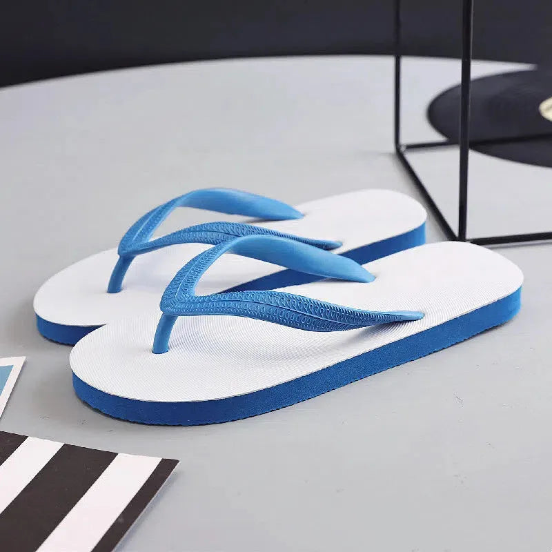A pair of rubber flip-flops with a fish logo on the strap, shown on a sandy beach.