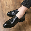 Men's black slip-on business casual shoes with tassel detail and plain toe on wooden floor