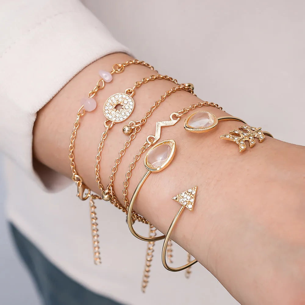 Set of six gold bracelets with arrow, V-shape, and water droplet charms on a dark background.