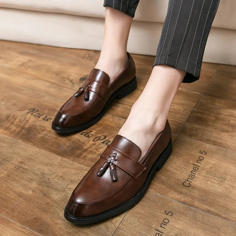 Men's black slip-on business casual shoes with tassel detail and plain toe on wooden floor