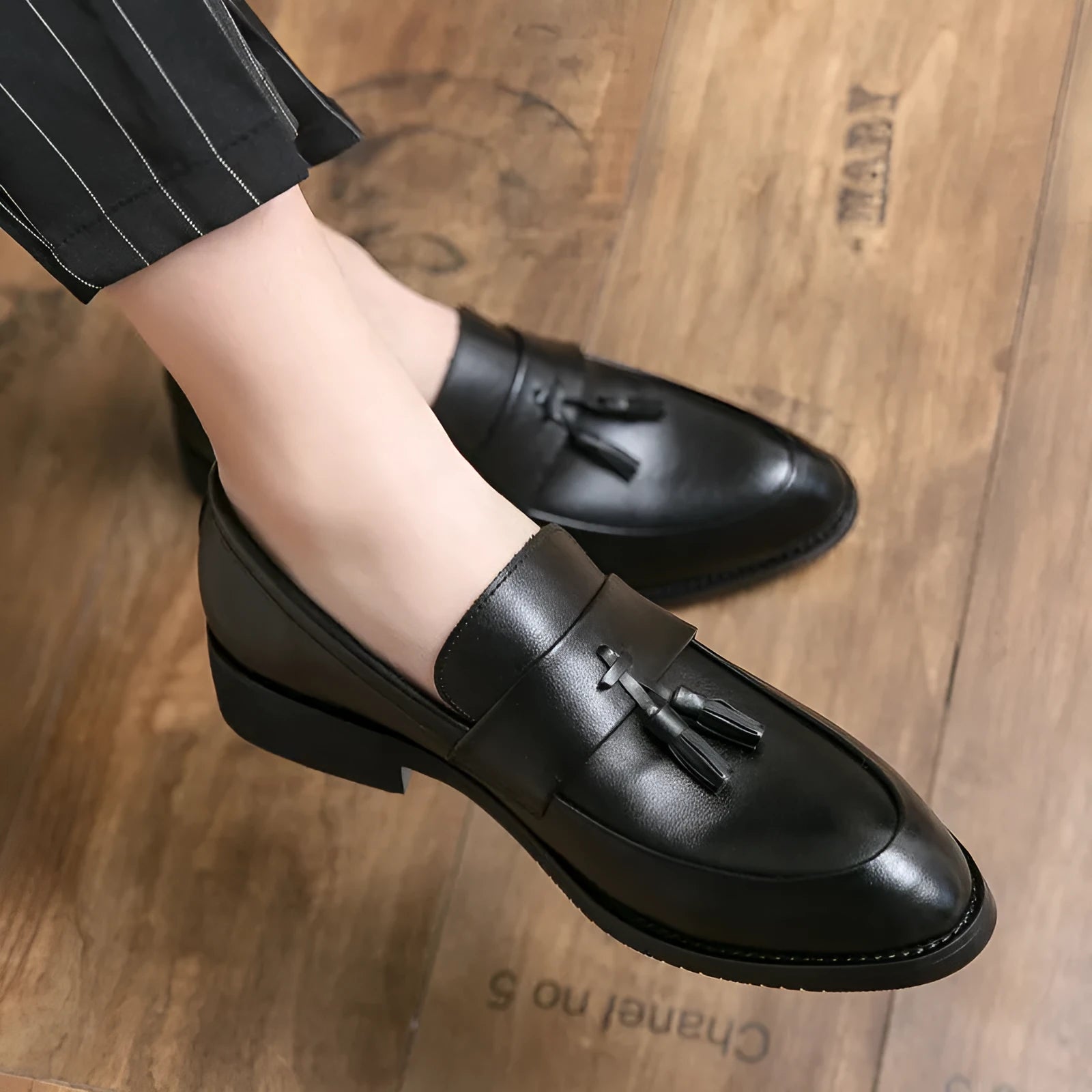Men's black slip-on business casual shoes with tassel detail and plain toe on wooden floor