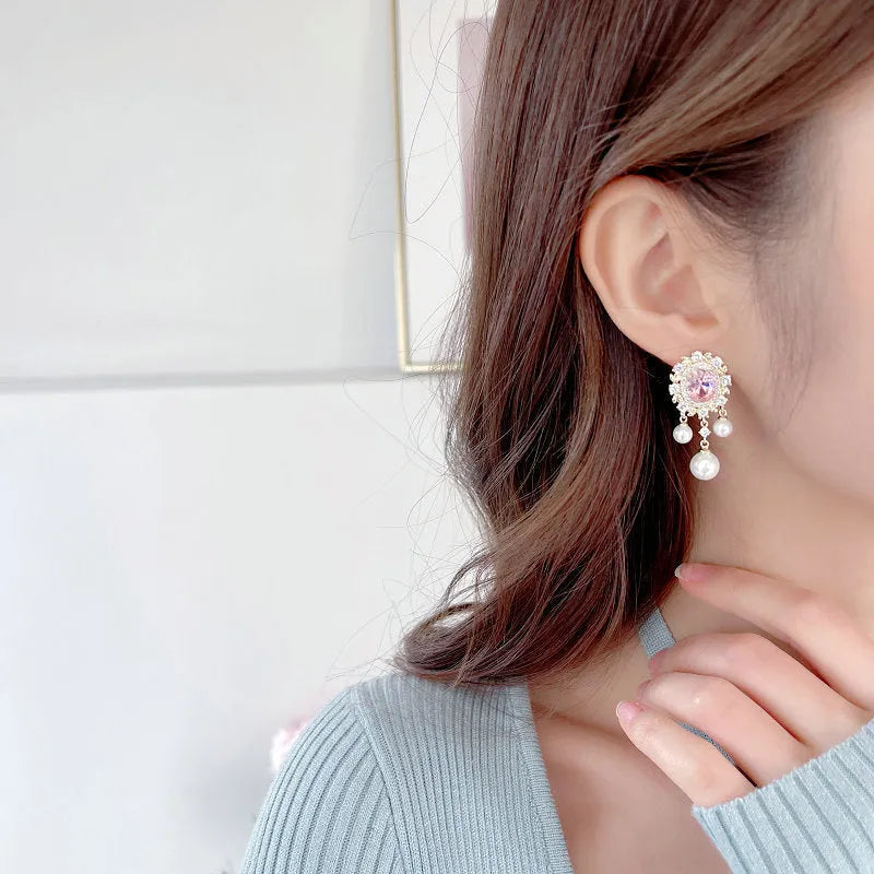 A pair of oval-shaped copper earrings with inlaid zircon stones and a dangling pearl tassel, shown in light purple.