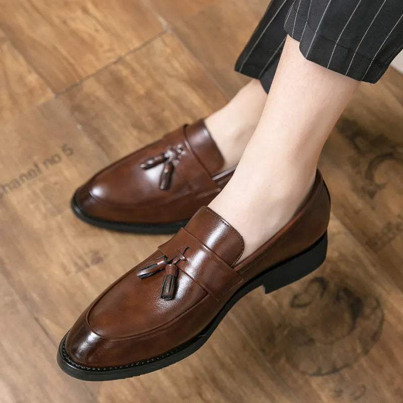 Men's black slip-on business casual shoes with tassel detail and plain toe on wooden floor